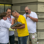 Mężczyzna w żółtym podkoszulku gratuluje stojącej na scenie kobiecie, obok mężczyzna i drugi mężczyzna mówiący do mikrofonu, za nimi elewacja budynku ratusza.