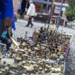 Zdobione, mosiężne flakoniki i inne przedmioty ułożone na ziemi, za nimi mężczyzna, oglądająca rzeczy kobieta.