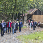 Kilkanaście osób idzie polną drogą, z tyłu las i drewniane zabudowania.