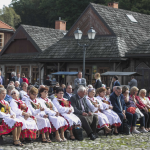 Grupa kobiet w strojach ludowych siedzi na drewnianych ławach, między nimi inne osoby, z tyłu drewniane zabudowania.