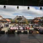 Starszy mężczyzna stojący tyłem na scenie, przed sceną tłum ludzi na ławkach, z tyłu drewniane zabudowania.