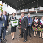 Stojące na scenie osoby: mężczyzna z kwiatkiem słonecznika, kobieta w mundurze i kobiety w strojach ludowych.