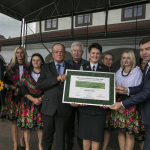 Grupa osób na scenie: kobiety w strojach ludowych, mężczyźni w garniturach, jeden z nich trzyma kwiat słonecznika.