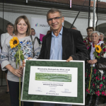 Kobieta i mężczyzna trzymają plakat z napisami, z tyłu kobiety w strojach ludowych.