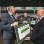 Mężczyzna w garniturze wręcza mężczyźnie plakat, z tyłu inni ludzie, wszyscy stoją na scenie.