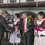 Mężczyzna gratuluje mężczyźnie trzymającemu kwiat słonecznika, obok kobiety w strojach ludowych.