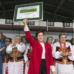 Stojąca na scenie kobieta w czerwonym żakiecie trzyma do góry plakat z napisami, za nią kobiety w strojach ludowych.