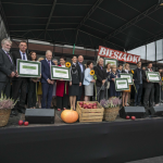 Grupa ludzi na scenie, niektórzy trzymają plakaty, na scenie drewniane skrzynki, owoce, wrzosy, z tyłu bilbord z napisem Biesiadka.