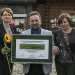 Mężczyzna trzyma plakat z napisami, po bokach dwie kobiety, z tyłu drewniane zabudowania.