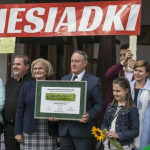 Grupa ludzi, mężczyzna trzyma biało-zieloną tabliczkę, u góry biały napis Biesiadki na czerwonym tle.