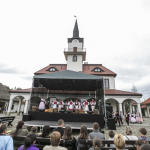 Budynek ratusza, przed nim plenerowa scena a na niej tańczące kobiety, przed nimi publiczność. Na scenie tańczące pary.