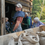 Kilka osób wchodzących do drewnianego budynku, obok drewniany wóz z workami ze zbożem.