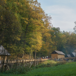 Drzewa w jesiennych barwach, drewniane zabudowania i ludzie idący mostkiem.