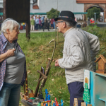 Stojący obok siebie starsza kobieta i mężczyzna, przed nimi kolorowe drewniane figurki, w oddaleniu ludzie.