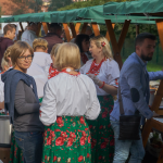 Ludzie przy drewnianych stoiskach z jedzeniem, kobiety w strojach ludowych.