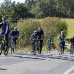 Grupa ludzi jadąca rowerami po asfaltowej drodze, z tyłu zarośla.