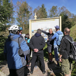 Kilkoro osób zgromadzonych przez mapą, jeden z mężczyzn na nią wskazuje, z tyłu drzewa na tle nieba.