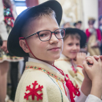 Chłopiec w okularach w stroju góralskim, za nim inny chłopiec i ludzie w przebraniach.