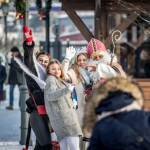 Dziewczynka przebrana za diabełka, dwie dziewczyny obok św. Mikołaj, z tyłu ludzie i drewniane budynki.