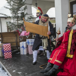 Mężczyzna w stroju króla siedzi na tronie, obok mężczyzna przebrany za Żyda odczytuje coś ze zwoju, z tyłu prezenty świerk i otwarta skrzynia.