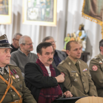 Mężczyzna w szaliku siedzi między mężczyznami w mundurach wojskowych, z tyłu inni ludzie