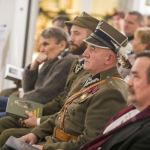 Siedzący na widowni mężczyźni, dwóch ma na sobie wojskowe mundury.