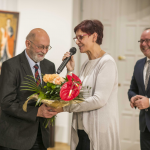 Kobieta wręcza starszemu mężczyźnie bukiet kwiatów, obok uśmiechnięty mężczyzna, na ścianie obraz.