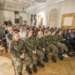 Siedząca na krzesłach publiczność, na pierwszym planie dziewczyny w wojskowych mundurach.