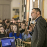 Mówiący do mikrofonu mężczyzna, przed nim laptop na stoliku, dalej publiczność na sali.