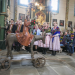 Figurka Chrystusa na osiołku, z tyłu ludzie w regionalnych strojach zgromadzeni we wnętrzu kościoła.