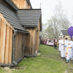 Lektorzy idący w procesji, niosący krzyż zakryty płótnem, z tyłu procesja, obok drewniany kościół.