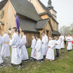 Lektorzy idący w procesji, niosący krzyż zakryty płótnem, z tyłu procesja, obok drewniany kościół.