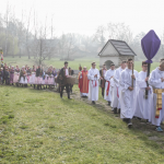 Lektorzy idący w procesji, niosący krzyż zakryty płótnem, z tyłu procesja, obok drewniany kościół.