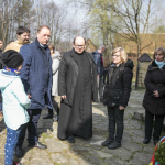 Trzy kobiety w strojach ludowych na łące, z tyłu drzewa i domy.