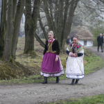 Dwie kobiety w strojach ludowych idące polną drogą, z tyłu ludzie i drzewa.