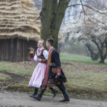 Dziewczyna i chłopak w strojach regionalnych idący polną drogą, z tyłu dom i drzewa.