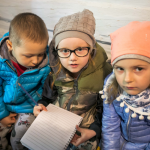 Dwie małe dziewczynki i chłopiec siedzący na ławce, jedna dziewczynka trzyma notes i ołówek.