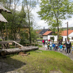 Idąca drogą grupa dzieci, kilkoro dorosłych, z tyłu budynki, drzewa, drewniane bale.