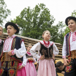 Dzieci w strojach ludowych stoją na drewnianej scenie, za nimi drewniane domy.