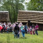 Grupa dzieci zgromadzona na trawie, z tyłu drewniane domy i drzewa.