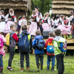 Grupa dzieci, dzieci w strojach ludowych tańczą na scenie, z tyłu drewniane domy.