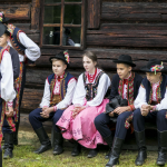 Dzieci w strojach regionalnych siedzą na ławce przed drewnianym domem, obok stoją chłopcy w strojach.