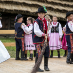 Dzieci w strojach ludowych tańczą na scenie, chłopiec z miotłą.