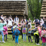 Dzieci w strojach ludowych na scenie, z tyłu drewniane domy, przed sceną grupa dzieci.