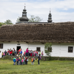 Drewniany, bielony dom, z tyłu widoczne wieże kościoła, przed domem duża grupa dzieci.