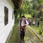 Chłopiec w stroju ludowym idący wzdłuż domu, z tyłu dzieci, dom i drzewa.