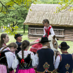 Dziewczynka w stroju regionalnym siedząca na koniu, za nią drewniany dom i drzewa, przed nią dzieci w strojach regionalnych.