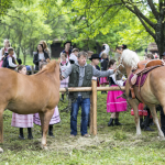 Dwa konie, między nimi mężczyzna, z tyłu dzieci w strojach regionalnych.