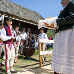 Grupa górali z instrumentami, z tyłu drewniany dom, na scenie kobieta z bochenkiem chleba.