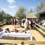 Stół, na nim gliniane naczynia, wokół niego ludzie w strojach ludowych, zgromadzona publiczność, z tyłu domy.